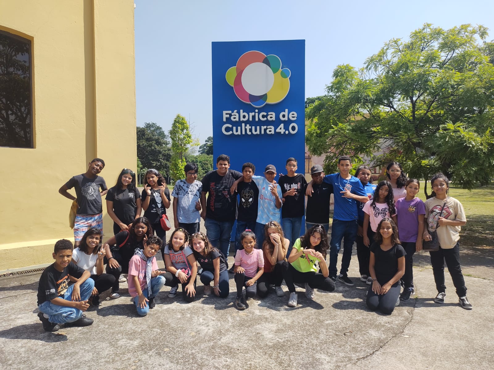 15 crianças estão em pé em frente a uma placa azul na qual se lê: Fábrica Cultural 4.0. Há, ainda, 10 crianças agachadas em frente de todos. No fundo, é possível ver uma parede amarela e diversas árvores. 